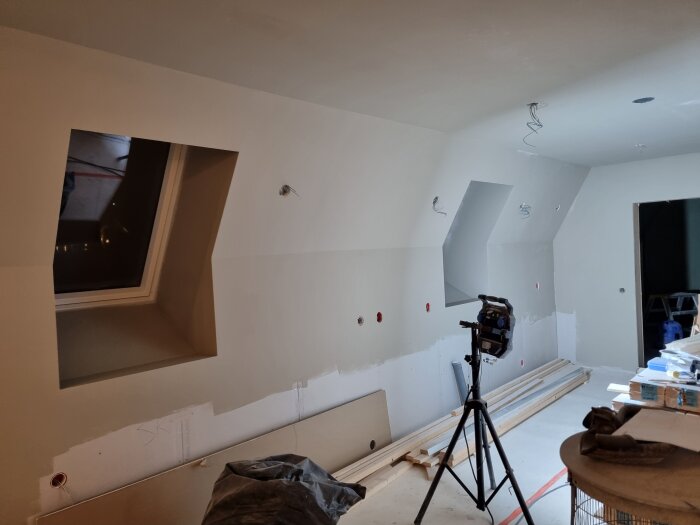 Rum under renovering med spacklade och grundmålade väggar, stora takfönster, träplankor på golvet, och en bygglampa på stativ.