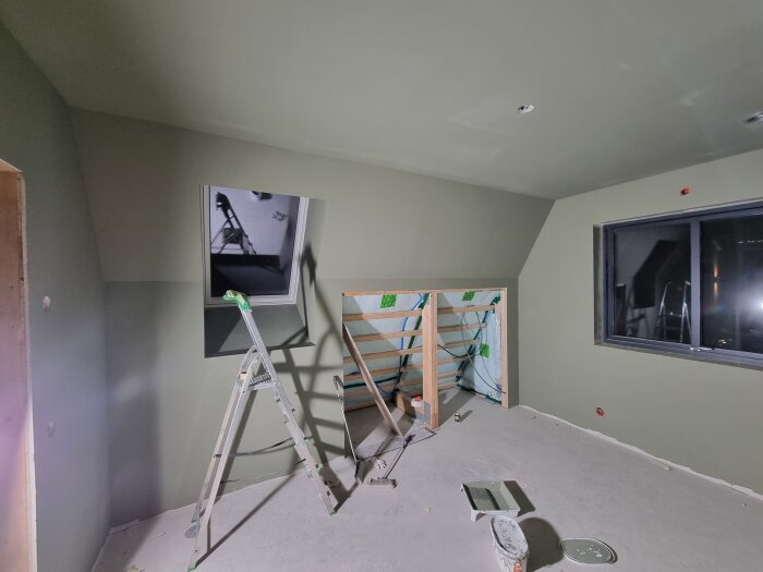 Rum under renovering med stege, nymålade väggar och golvtillbehör. Fönster till höger och ett utrymme för rör- och elinstallationer syns.