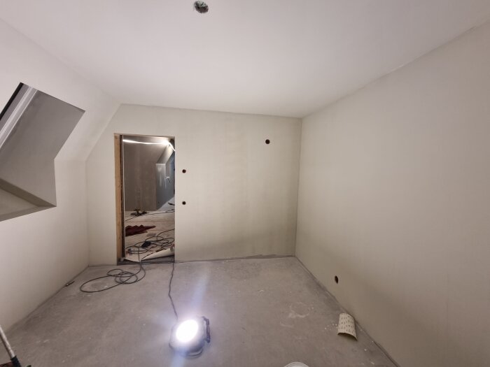 Rum under renovering med spacklade väggar och tak, betonggolv och arbetslampa på golvet. Kablar och verktyg syns i bakgrunden genom en dörröppning.
