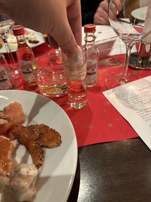 Person doppar påse i glas med alkohol, omgiven av små flaskor och tallrik med lax på röd duk.