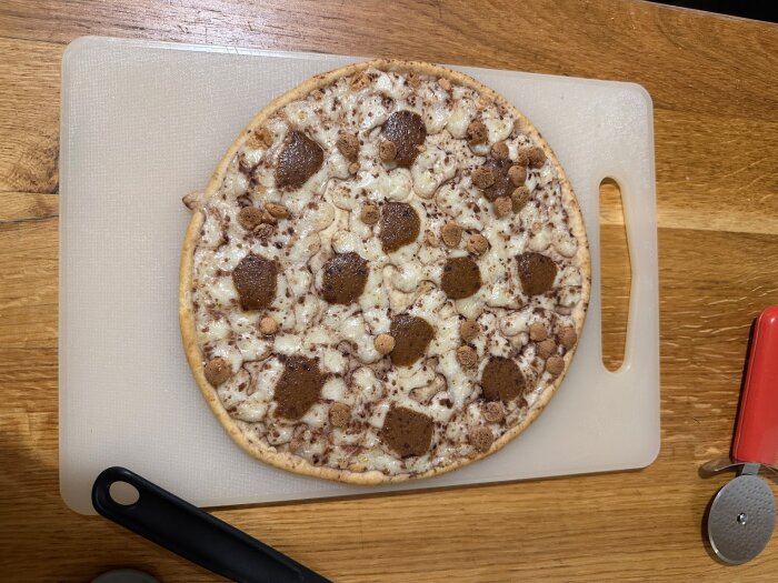 Dessert-pizza på skärbräda, ser ut som tiramisu med mörka fläckar och strössel, på ett träbord.