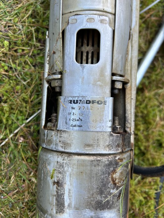 Trasig vattenpump i rostfritt stål med en etikett som visar tekniska specifikationer, liggande på gräsmatta.