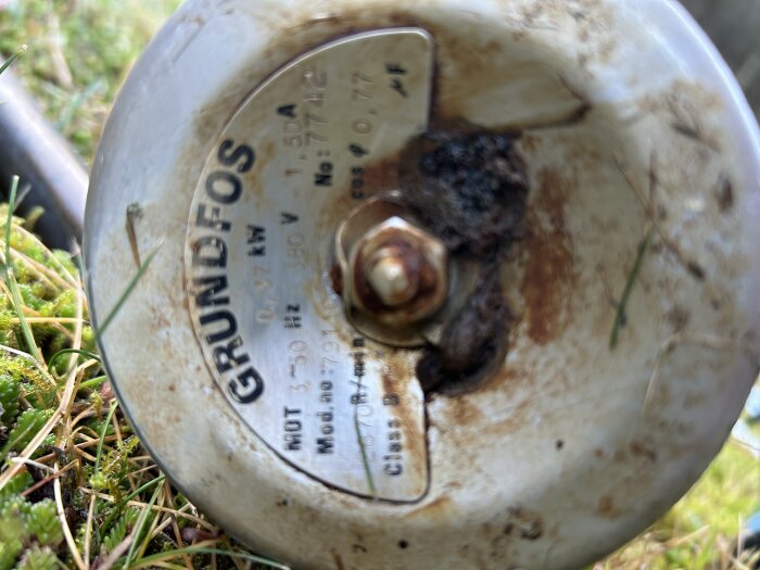 Rostig Grundfos vattenpump i närbild, med etikett synlig, liggande i gräs. Modellspecifikationer delvisa.