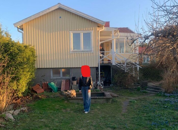 Gul träfasad på ett äldre hus från 1936, med hög sockelvåning och rymlig trädgård. Solnedgångsbelyst, med person i förgrunden.