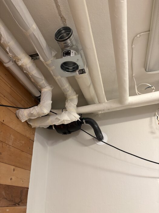 T-rör för ventilation monterat i tak, med nedåtriktad pax-fläkt och omgivande isolerade rör. Träpanel och vitmålade väggar i bakgrunden.