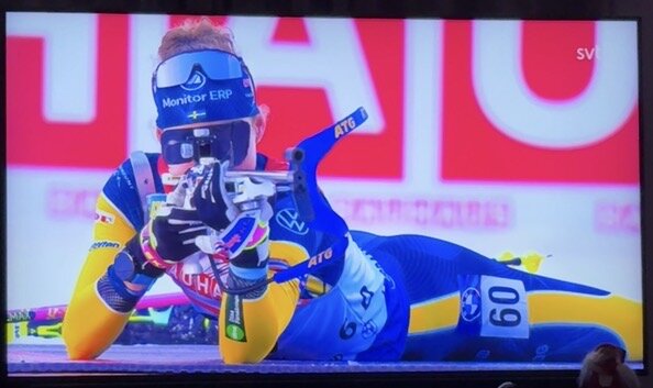 En skidskytt i gul-blå dräkt ligger i skjutposition med gevär under en tävling.