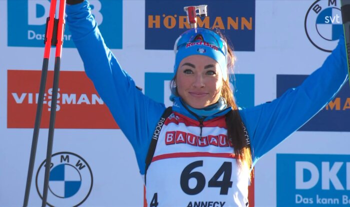 Kvinna i blå skidutrustning med startnummer 64 firar med armarna i luften framför sponsorvägg vid en skidtävling.