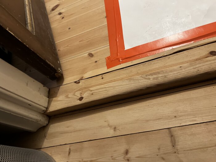 Trätröskel på ett plankgolv vid en dörr, med vit papperstätning upptejpad med orange tejp i hörnet.