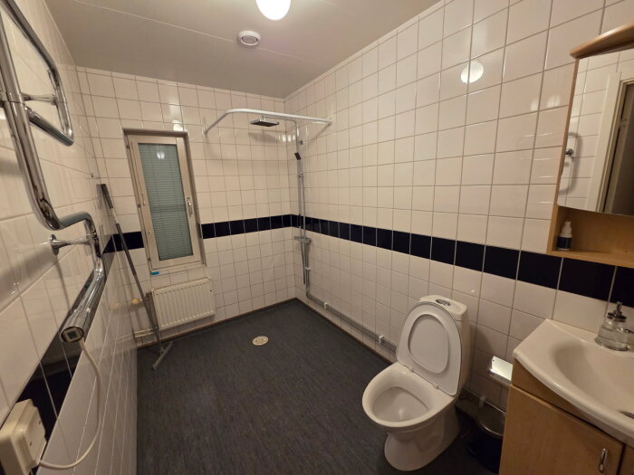 Badrum med vita kakelväggar och grå golvmatta, toalett, handfat och duschutrymme med rör. Möjlig fuktskada och renoveringsplaner nämns.