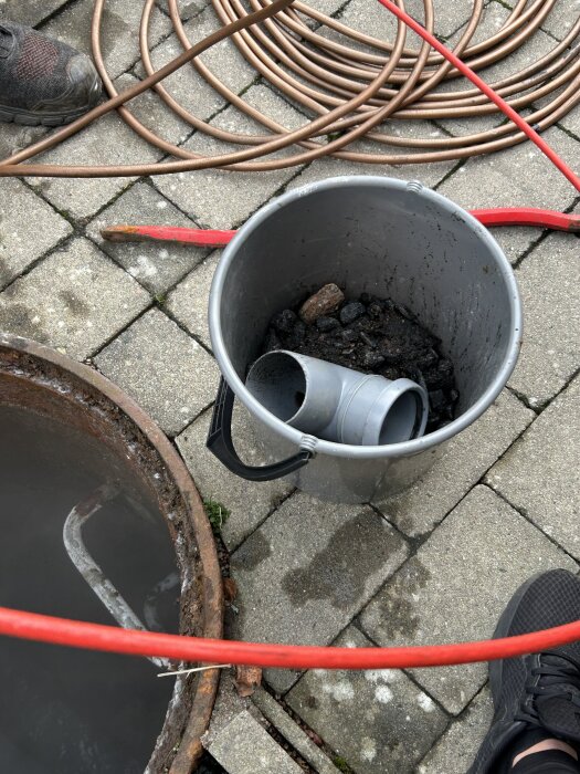 En grå hink fylld med stenar och ett grått rör ligger på marken bredvid ett öppet avloppsbrunn, omgiven av slangar och en skosula.