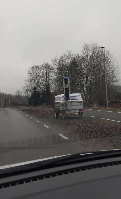 Släpvagn med fartkamera placerad vid vägkant under molnig himmel med träd i bakgrunden.