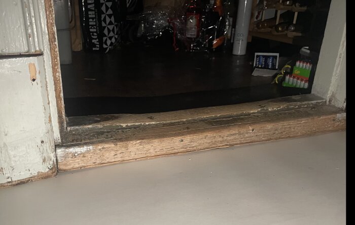 Gammal tröskel i slitet trä, troligen kärnfuru, med avflagnad färg och tecken på långvarigt användande i dörröppning.