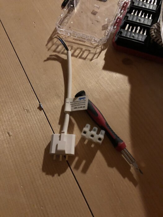 En vit kabel med trippelanslutning, en skruvmejsel och en genomskinlig plastlåda på träbord.