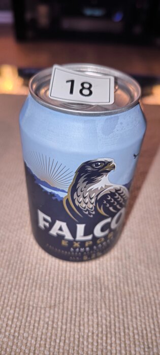 Kapsyl av en Falcon Export ölburk med en etikett som visar siffran 18 placerad ovanpå. Burkens design har en bild på en falk.