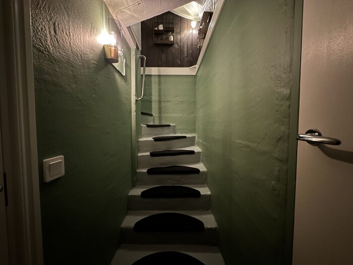 Grön målad källartrappa med ljus ovanför, tydligt renoverad med mörka stegskydd och modern belysning.