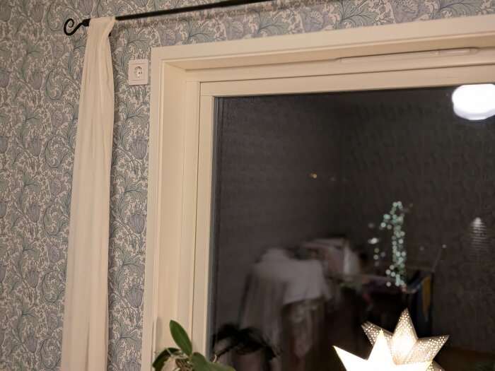 En vit gardin hänger vid ett fönster med ett eluttag placerat ovanför gardinstången på en mönstrad tapet.