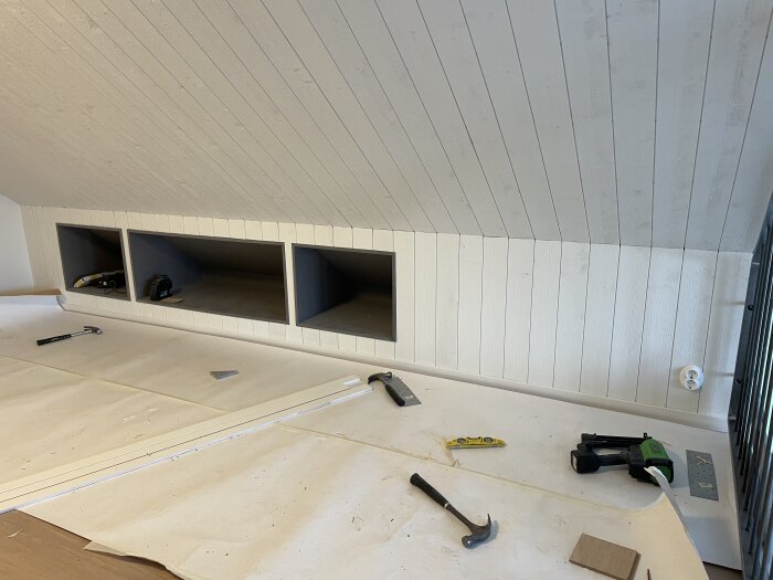 Byggprojekt i loft: panel på kattvinden klar, verktyg framför inkl. hammare och såg, samt påbörjat arbete med lister och ramar.