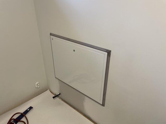 Vitt skåp för vattenfördelaren på vägg med ram av MDF, rör synligt. Primer på golvet och verktyg syns också.