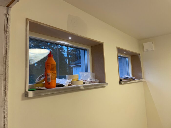 Fönsterfoder med spotlights ovanför, diverse byggmaterial och rengöringsprodukter på fönsterbrädan, synliga detaljer av pågående renovering.