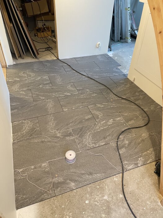 Nylagt grått klinkergolv i hall, löpande snöre från en spole och en elkabel på golvet, dörröppning med verktyg i bakgrunden.