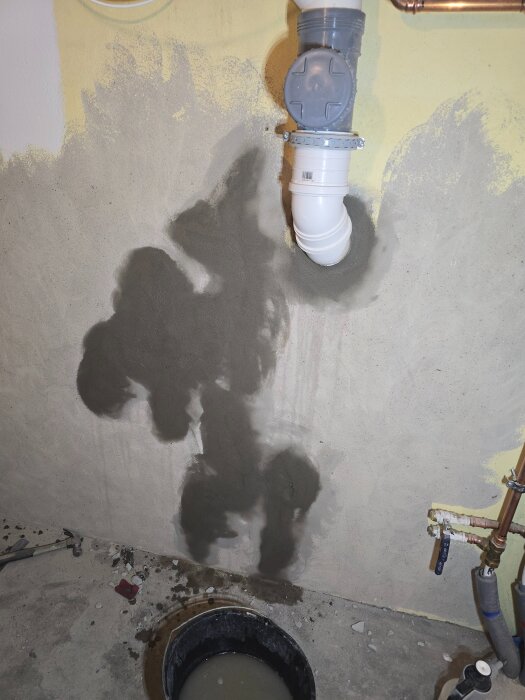 Vägg med nyligen lagade sprickor vid röranslutningar, intill en hink med vatten och en hammare på golvet i en pågående renovering.