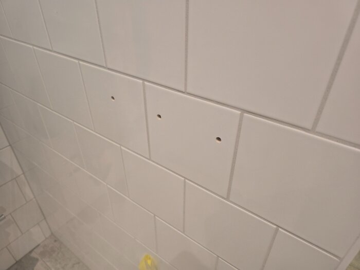 Närbild på kakelvägg med två hål förberett för montering av badrumstillbehör som handdukskrokar eller toalettpappershållare.