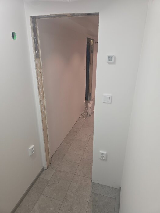 Vitmålad källargång med nylagt grått kakelgolv, eluttag på väggarna och synliga arbeten runt dörrkarmen.