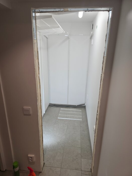 Renoverad klädkammare med vita väggar och golvplattor, synlig eluttag och ventilation, sedd genom en dörröppning utan dörrkarm.