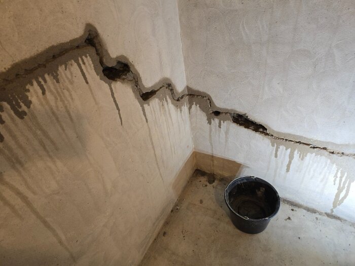 Sprickor i en källarvägg med delvis applicerat bruk syns vid en hörna; en svart hink står på betonggolvet.