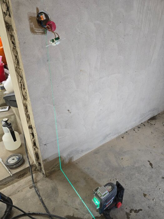 Laserverktyg projicerar en grön linje för kabeldragning på en betongvägg i ett renoveringsprojekt. Synliga verktyg och material vid sidan.