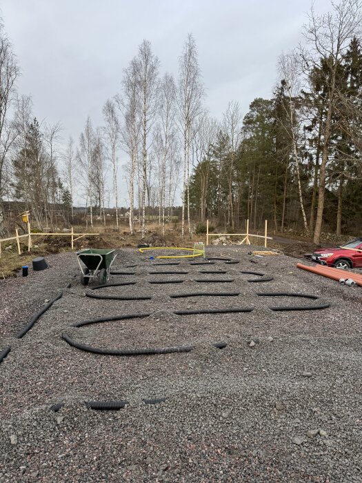 Byggarbetsplats med packad grusgrund och svarta rör lagda på marken, omgiven av träd och med en röd bil i bakgrunden.
