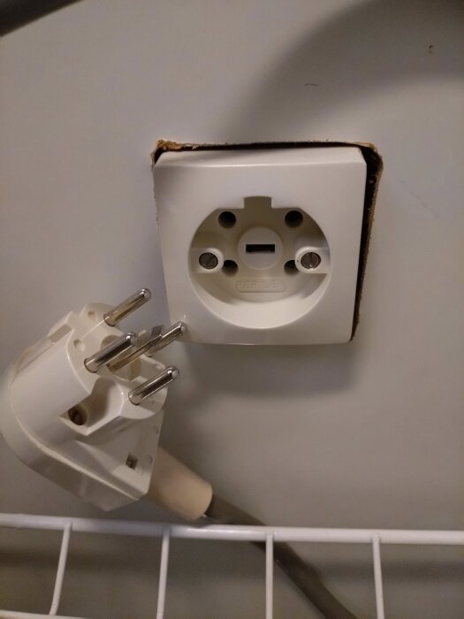 Vitt eluttag och stickpropp med tre stift, monterade i en vit vägg ovanför en metallhylla.