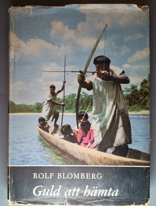 Omslag av Rolf Blombergs bok "Guld att hämta" med personer i en kanot på en flod, en man siktar med en pilbåge.