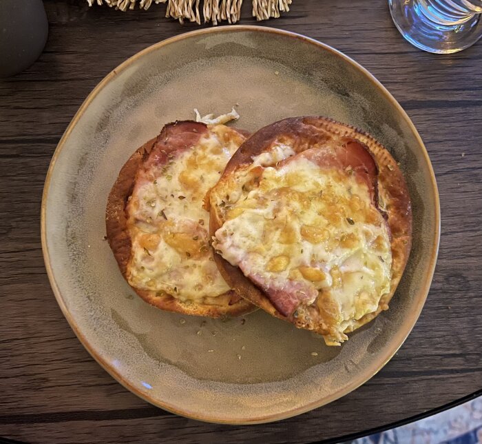 Två krispiga mini-pizzor med skinka och smält ost på en beige tallrik, placerade på ett mörkt träbord.