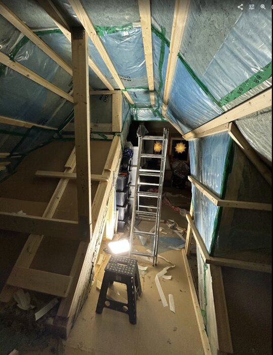 Vind med isolering, ångspärr och träreglar synliga. Stege och arbetsbelysning på golv. Arbetet är delvis klart, med oorganiserat byggmaterial.