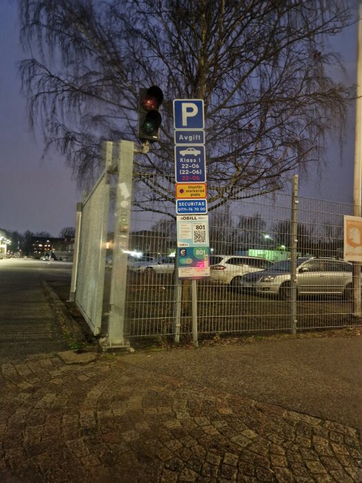 Parkeringsskylt med avgiftstider, trafikljus, parkering i bakgrunden, och stängsel vid en upplyst parkeringsplats i skymning.