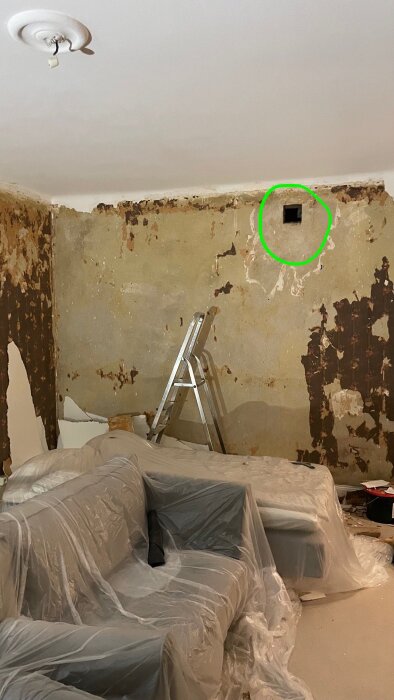 Renoverat rum med avskalat tapet, stege, täckt soffa och öppning i väggen som kan vara en ventilation eller gammal kakelugn.