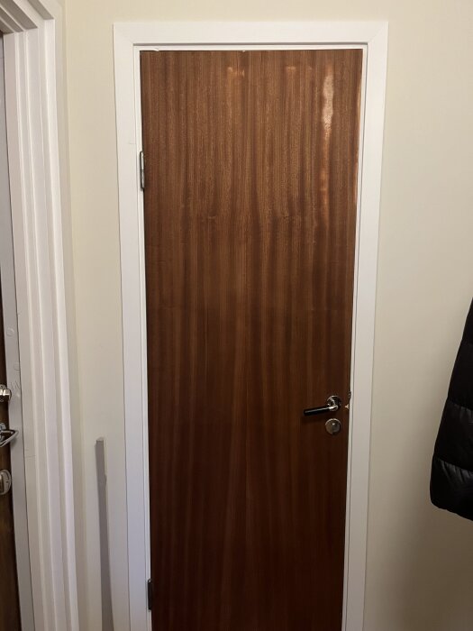 Trädörr med slipade märken och synliga skador i lacken, omgiven av vit karm och del av vägg.