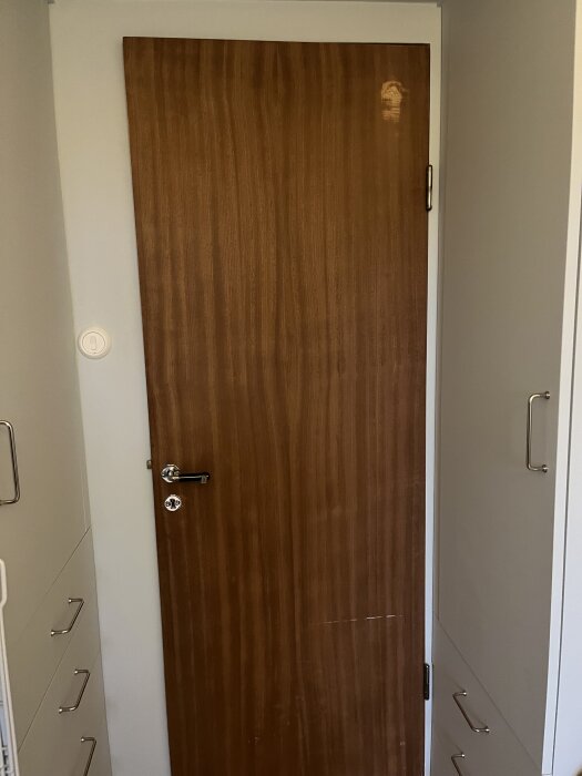 Trädörr med märken efter slipning, belägen mellan två vita väggar med handtag synliga.