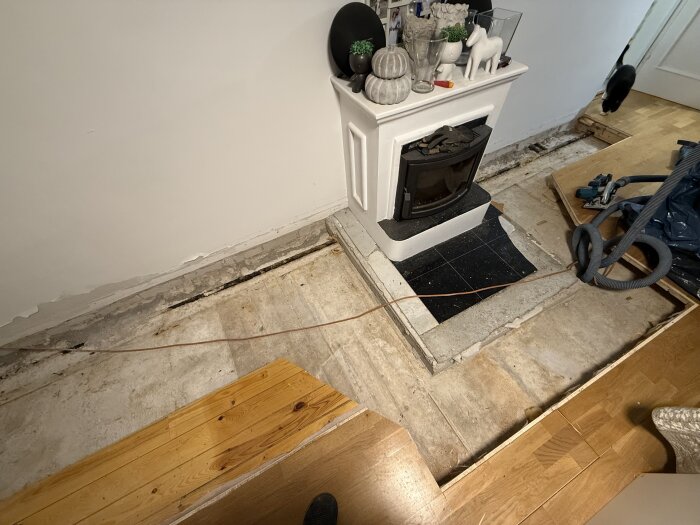 Brädgolv under renovering med framtagen plats för elementrör, framför en vit kamin dekorerad med prydnadssaker, och en katt i bakgrunden.