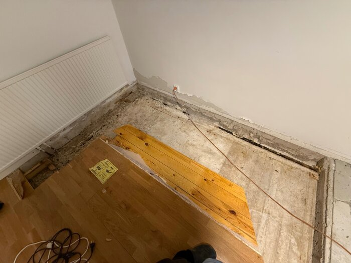 Ett rum under renovering med borttaget golv nära ett element, förberedelser för att installera nya elementrör syns. Olika golvmaterial avslöjas.