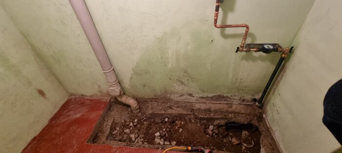Stort hål i golvet där huvudledningarna går in och ut i ett litet rum, med synliga rör och en mätare på väggen.