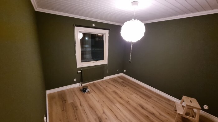 Sovrum under renovering med målade gröna väggar, trälaminatgolv och vit taklampa. Färgburk och pall på golvet.