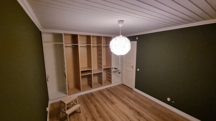 Rum med gröna väggar och träparkett, garderob under montering och vit taklampa i fokus. Trästegpall står framför garderoben.