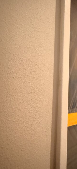 Närbild av ett vägg- och dörrkarmshörn med synliga målarlinjer och penseldrag, delvis täckt av plastskydd med gul tejp.