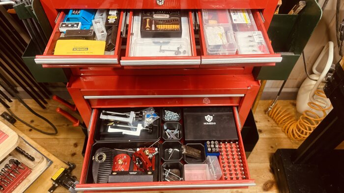 Röd verktygsvagn med öppna lådor fyllda med organiserade verktyg, bits och tillbehör, inklusive metallplattor och förvaringslådor.