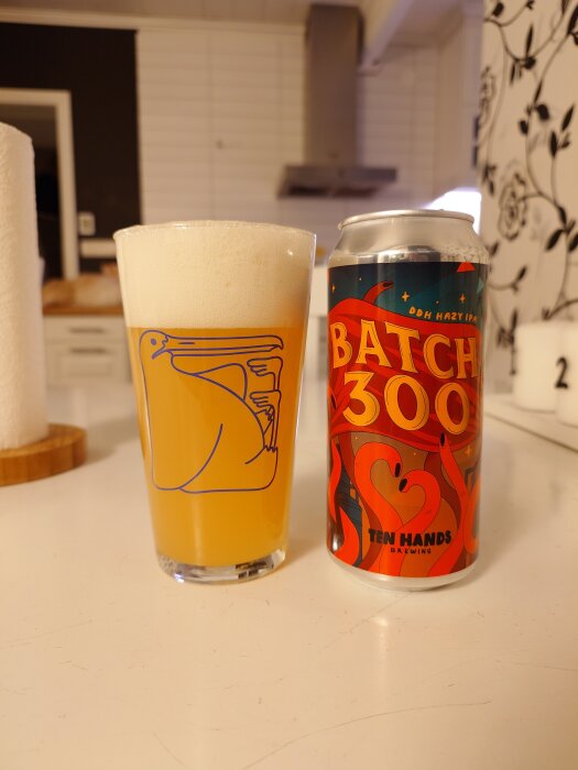 Ölglas med skummande öl bredvid en burk "Batch 300 Ten Hands", en DDH Hazy IPA, på ett köksbord.