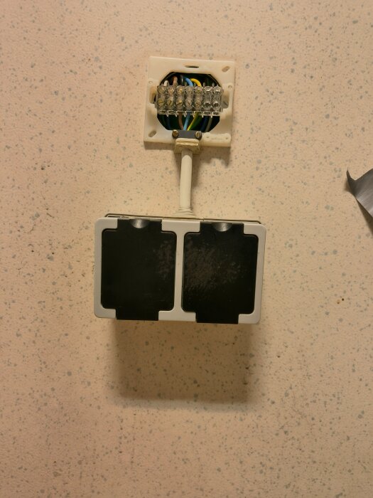 Dubbel väggkontakt monterad på ett öppet uttag med synliga kablar och kopplingsblock, installerat på en ljus vägg.