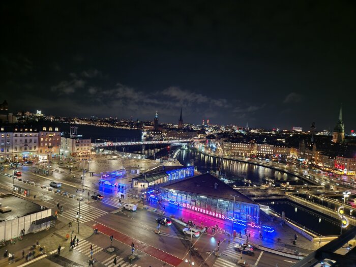 Nattlig vy över en upplyst stad med bilar, broar och byggnader vid vattnet, där neonskyltar och ljus reflekteras i reflekterande ytor.