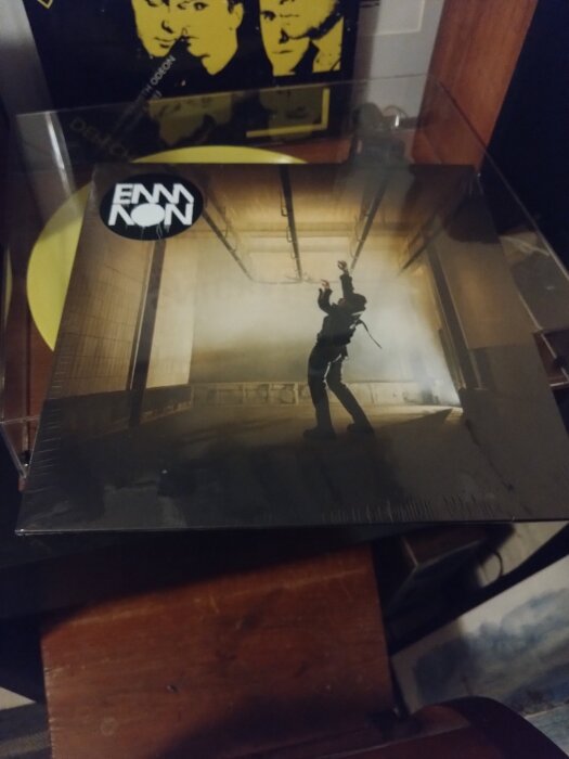 Omslag av album från bandet EMMON, med motiv av en person som sträcker sig uppåt i en industriell miljö, stående på ett golv med metallrör i taket.